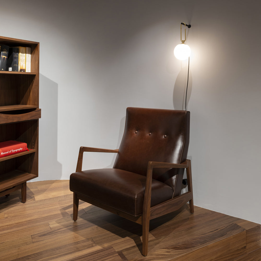 Small and lit in the corner of a grey room, a Clérachic globe wall lamp hangs above a leather armchair and a walnut bookcase.