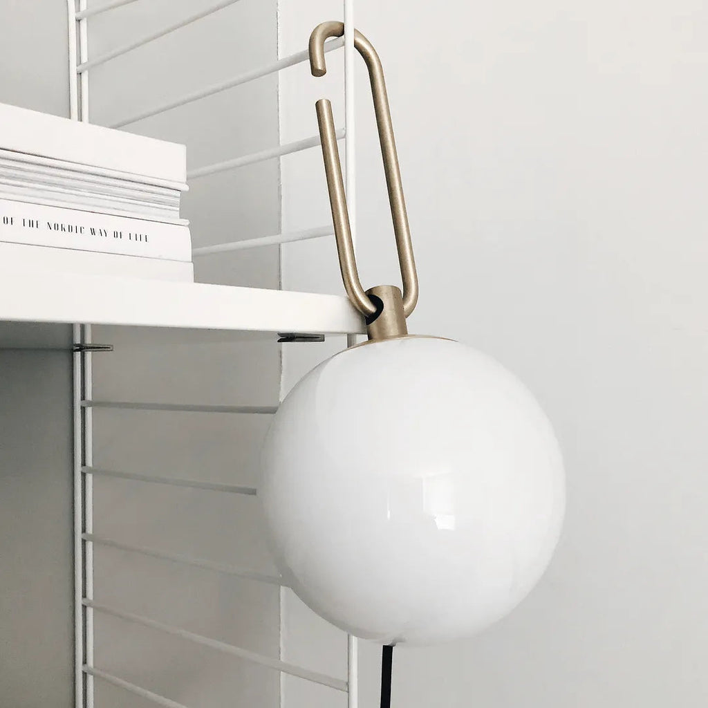 The gold hook of this Clérachic globe lamp hooked over a white shelving system, with books stacked on the shelf beside it.
