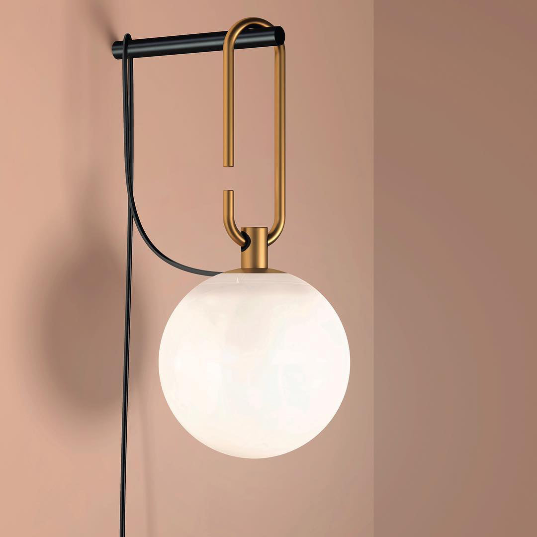 A white globe lamp from Clérachic on a gold loop and black bar, against a soft pink-beige wall with its cord trailing down.