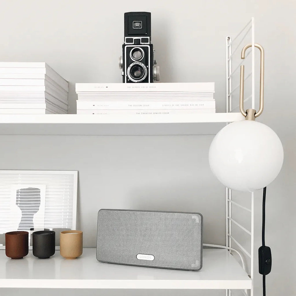A Clérachic globe lamp hangs from a white shelf above a speaker, ceramic cups and stacked books, its cord running down to an inline switch.