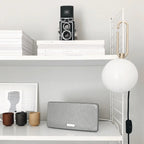 A Clérachic globe lamp hangs from a white shelf above a speaker, ceramic cups and stacked books, its cord running down to an inline switch.