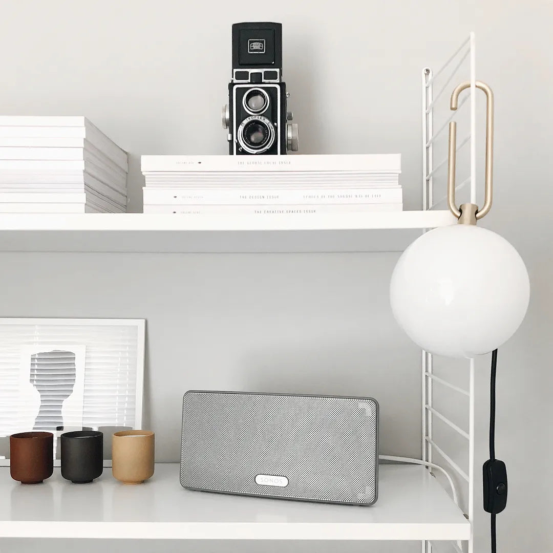A Clérachic globe lamp hangs from a white shelf above a speaker, ceramic cups and stacked books, its cord running down to an inline switch.
