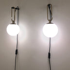 Two Clérachic globe wall lamps lit cool white on a pale wall, each with a gold loop and a black cord with an inline switch.
