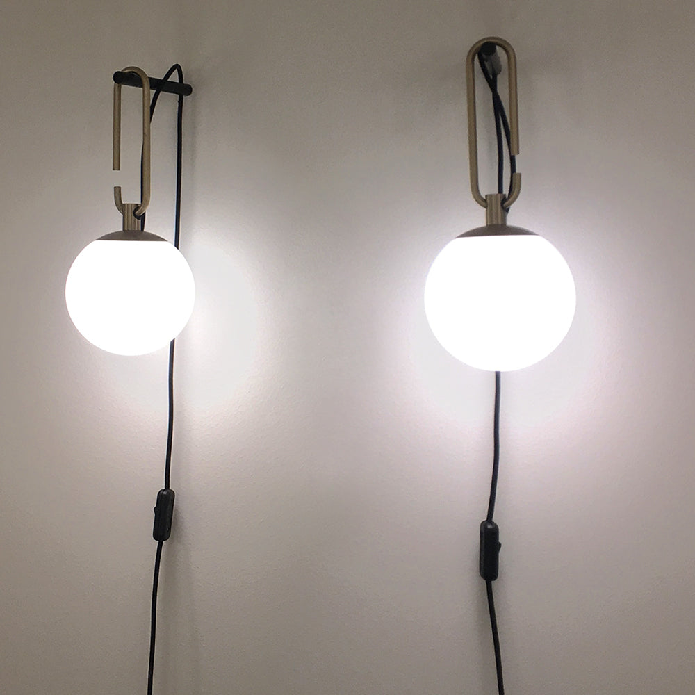Two Clérachic globe wall lamps lit cool white on a pale wall, each with a gold loop and a black cord with an inline switch.