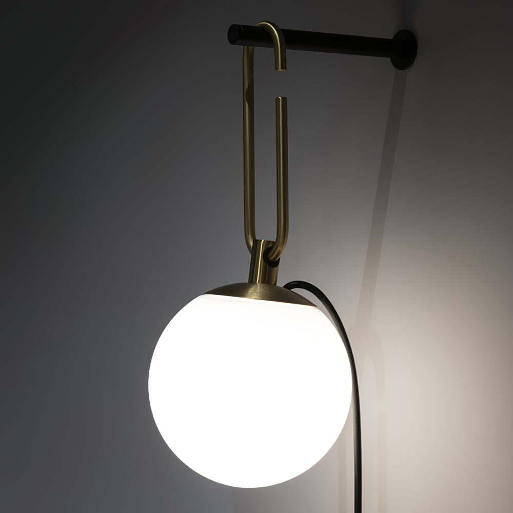 A Clérachic globe lamp lit white against a dark grey wall, hung from a gold loop on a black bar.