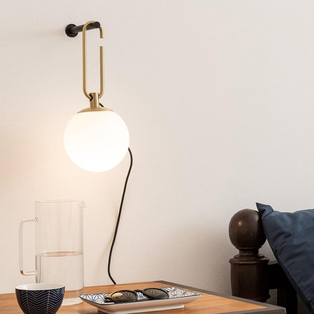Close view of a Clérachic globe wall lamp on a pale wall above a glass jug, a bowl and sunglasses on a wooden side table.