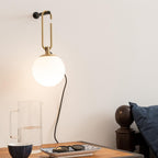 Close view of a Clérachic globe wall lamp on a pale wall above a glass jug, a bowl and sunglasses on a wooden side table.
