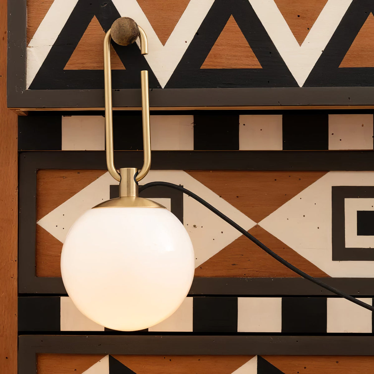 A Clérachic globe lamp lit white, hanging from a wooden knob in front of a chest painted with black, white and rust triangles.