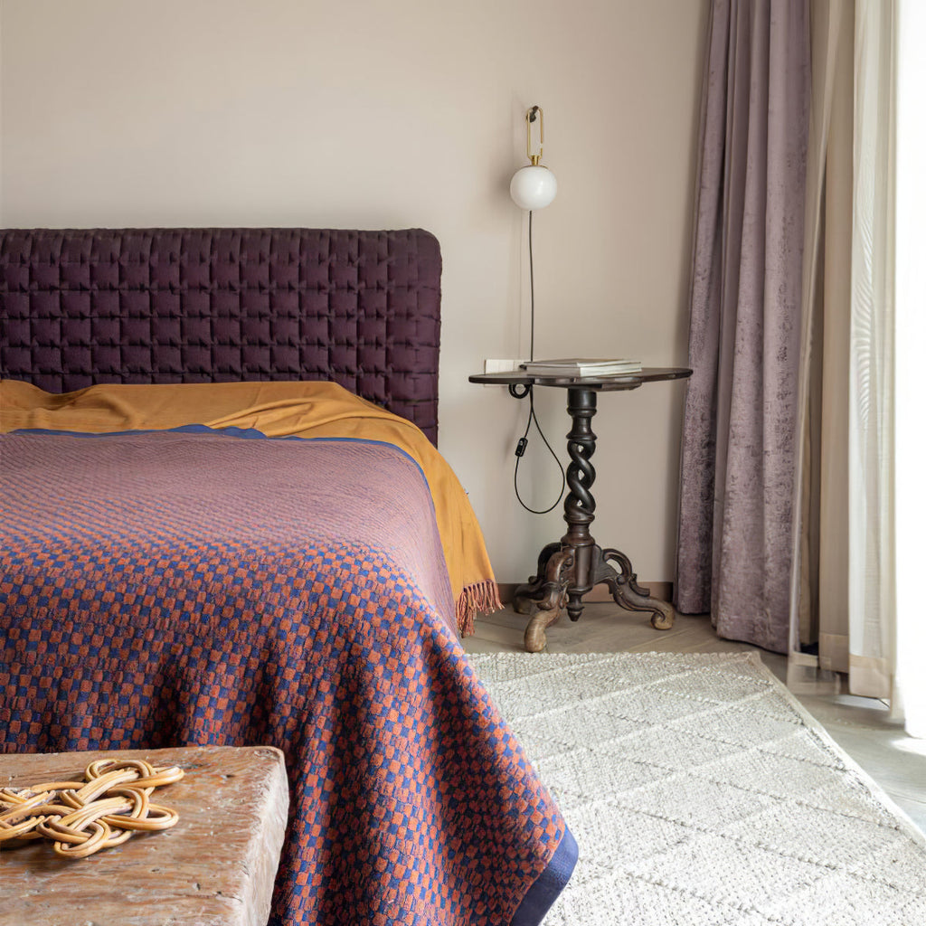 A small Clérachic globe wall lamp on a pale wall above a bed with a plum headboard, beside a lilac curtain and a carved side table.