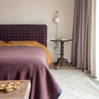 A small Clérachic globe wall lamp on a pale wall above a bed with a plum headboard, beside a lilac curtain and a carved side table.