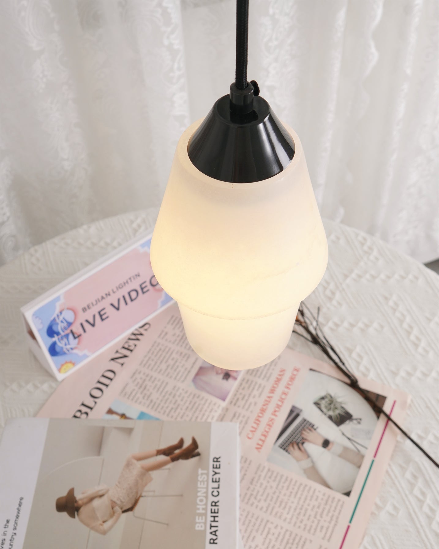 Clérachic Black and White Alabaster Pendant Light glowing over a lace tablecloth with a newspaper and dried stems.