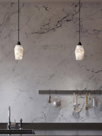 Two Clérachic Black and White Alabaster Pendant Lights hang over a marble kitchen wall with a black counter and faucet.