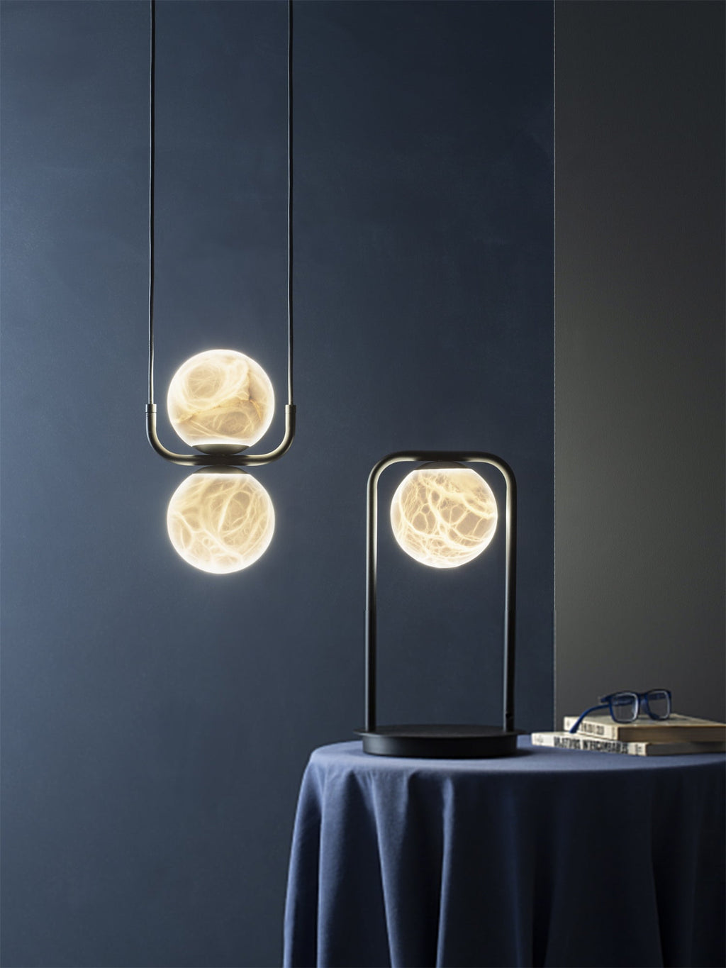 The Clérachic two-globe pendant against a deep blue wall, veining glowing through the stone beside a lit table lamp.
