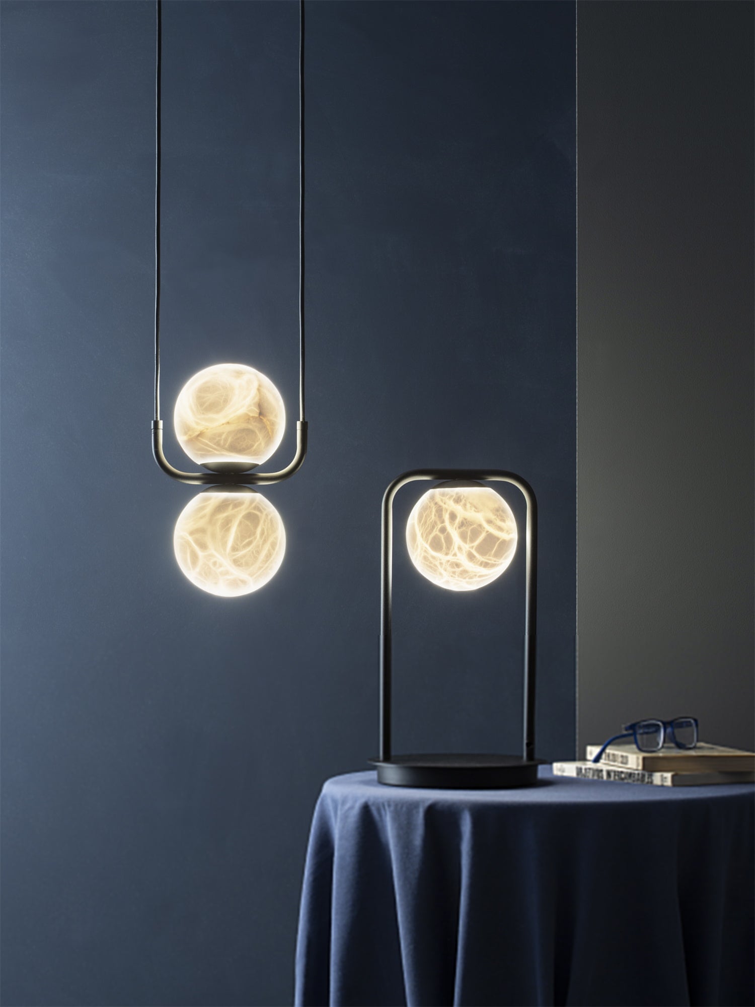 The Clérachic two-globe pendant against a deep blue wall, veining glowing through the stone beside a lit table lamp.