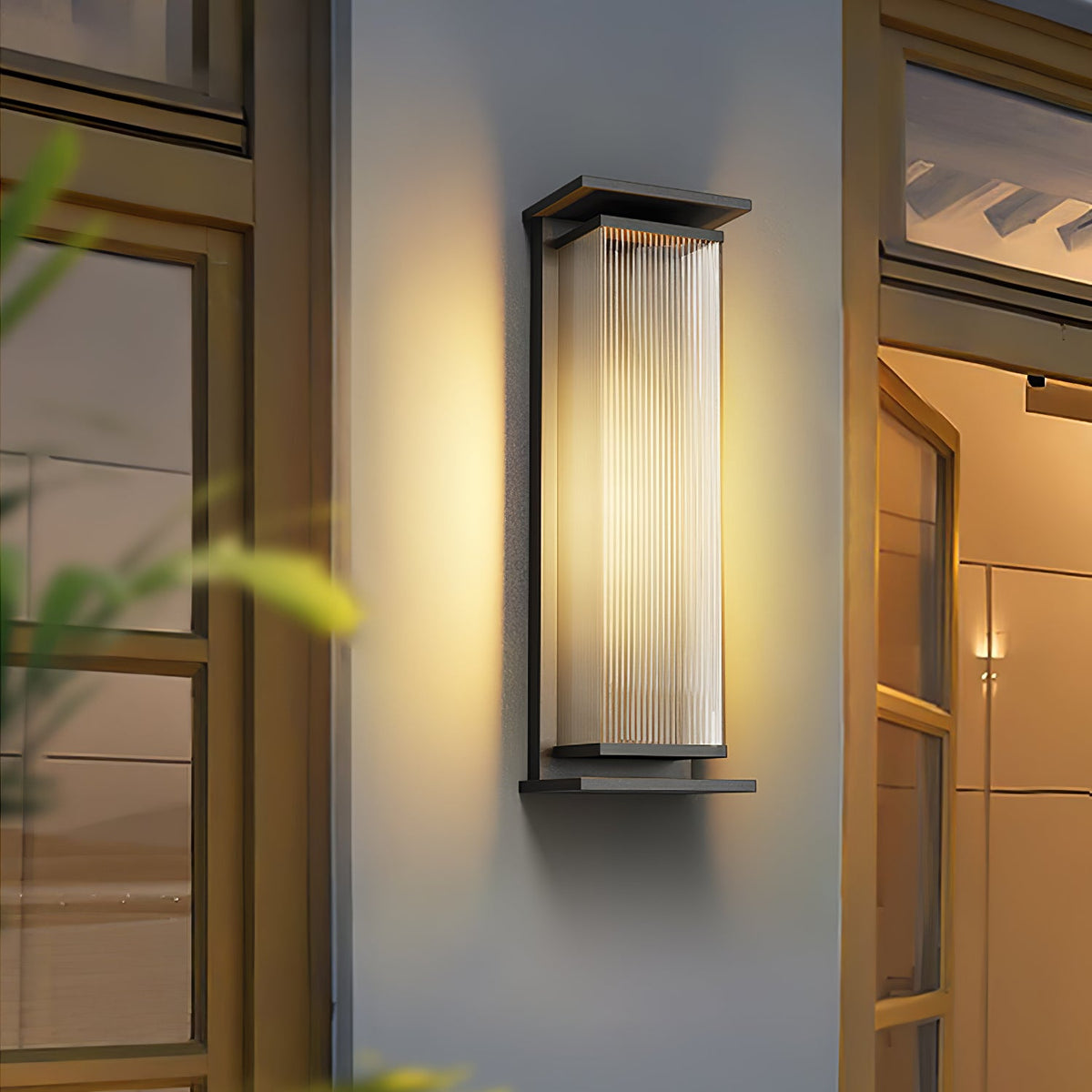 A lit Clérachic wall light beside a glazed door on a pale rendered wall, its black frame and ribbed shade casting a warm halo.