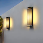 Two Clérachic wall lights in different heights on a pale outdoor wall at dusk, both glowing warm against a blue sky.