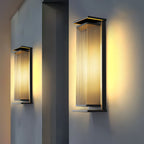 A tall and a shorter Clérachic wall light side by side on a grey wall, beside a dark column, both casting warm light.