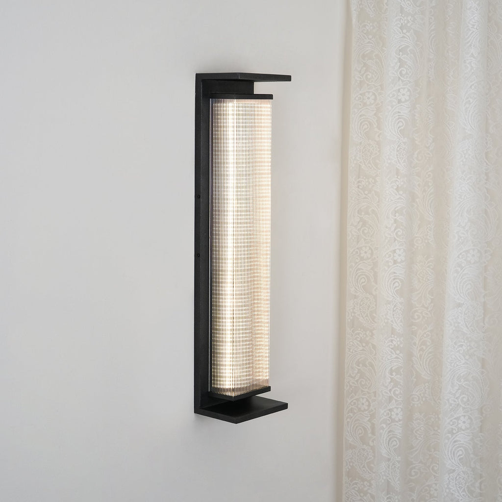 Side view of a Clérachic wall light in a black frame, with a ribbed shade lit white against a pale wall and a lace curtain.