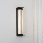 This Clérachic wall light lit cool white, mounted on a pale wall beside a lace curtain, with its black frame and shelf-like base.