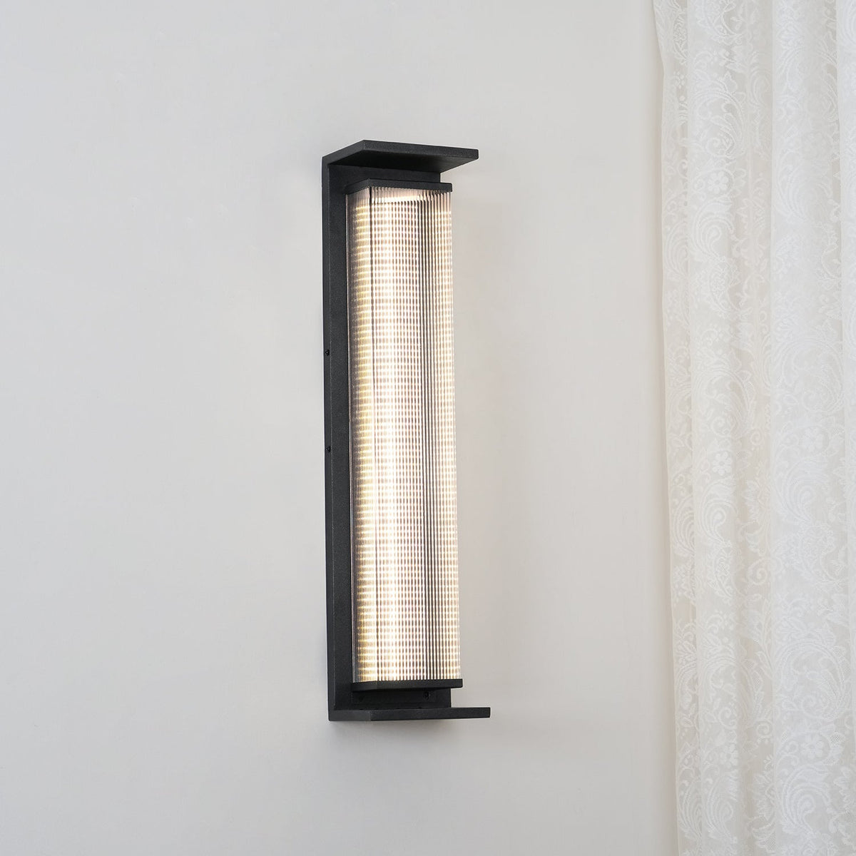 This Clérachic wall light lit cool white, mounted on a pale wall beside a lace curtain, with its black frame and shelf-like base.