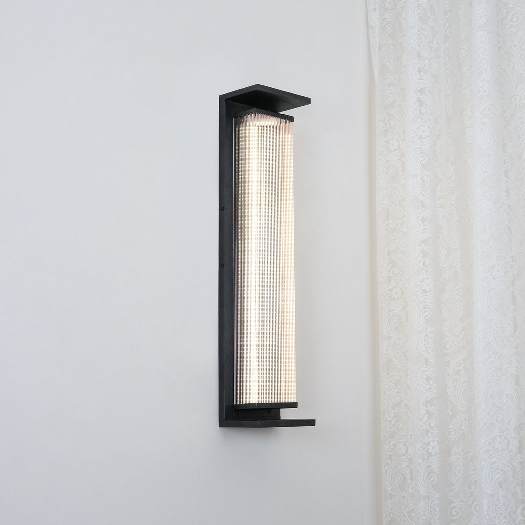 A Clérachic wall light shown from a slight angle, its ribbed shade glowing white inside a black frame, beside a lace curtain.
