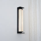 A Clérachic wall light shown from a slight angle, its ribbed shade glowing white inside a black frame, beside a lace curtain.