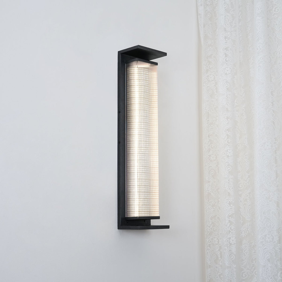A Clérachic wall light shown from a slight angle, its ribbed shade glowing white inside a black frame, beside a lace curtain.