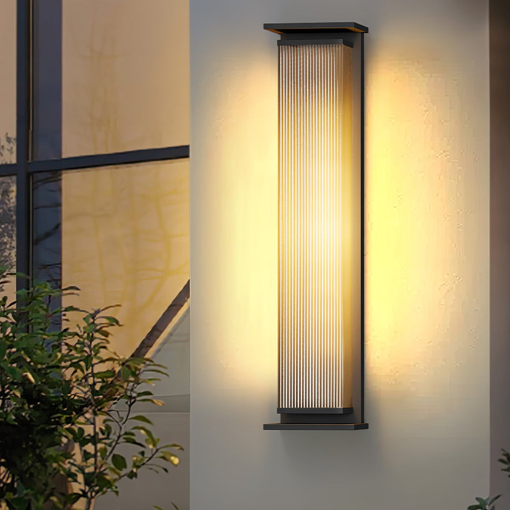 A tall Clérachic wall light beside a window with a leafy shrub below, its ribbed glass shade lit warm.