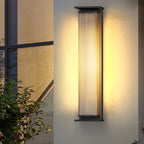 A tall Clérachic wall light beside a window with a leafy shrub below, its ribbed glass shade lit warm.