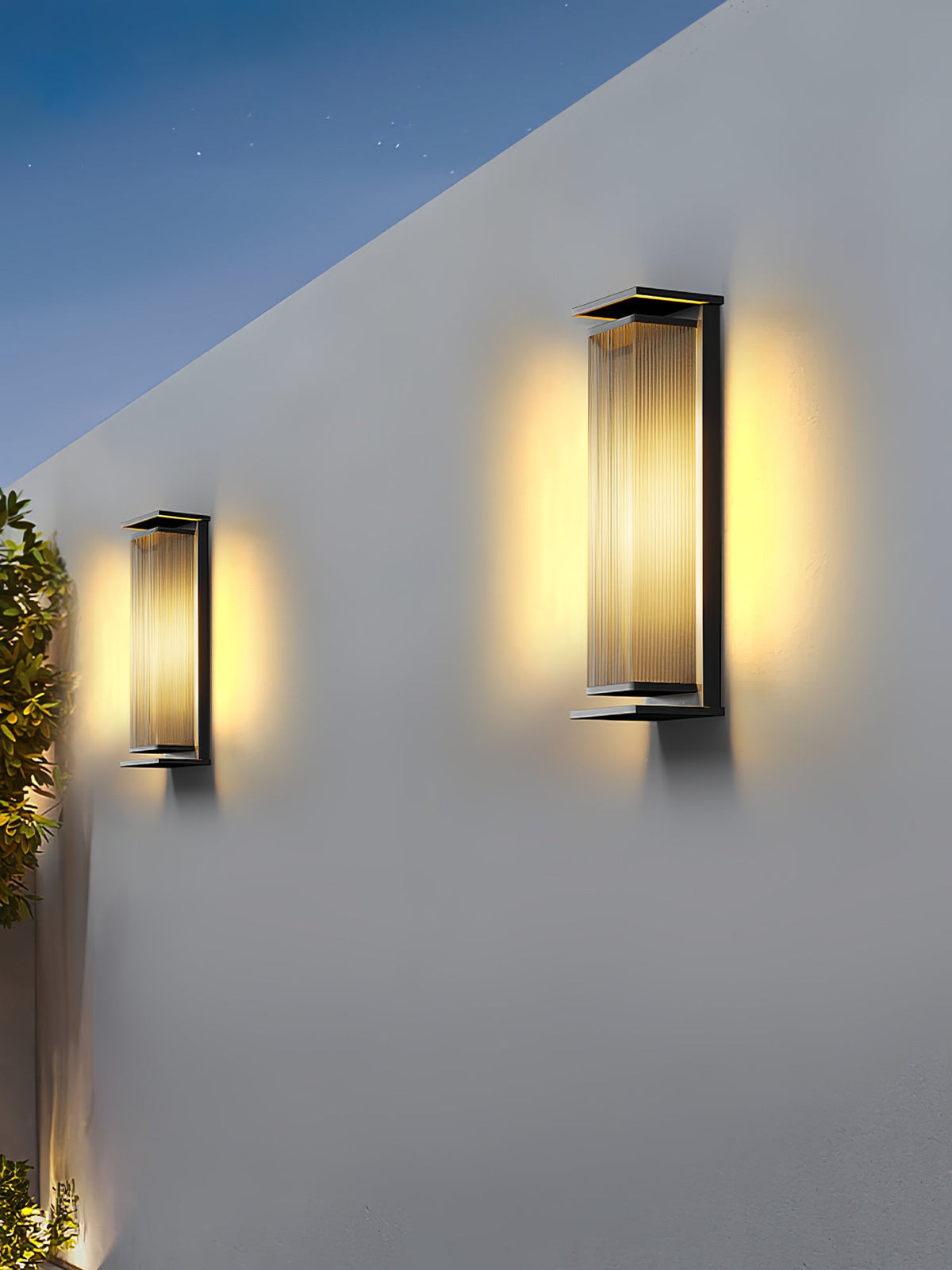 Two Clérachic wall lights of different heights on a white wall at dusk, their ribbed shades glowing warm under a darkening sky.
