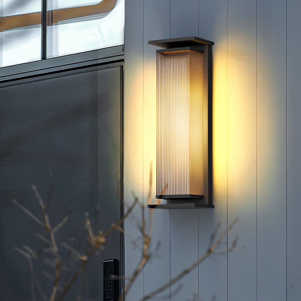 A Clérachic wall light on a pale cladded wall by a dark door, its ribbed shade lit warm against bare branches.
