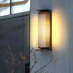 A Clérachic wall light on a pale cladded wall by a dark door, its ribbed shade lit warm against bare branches.