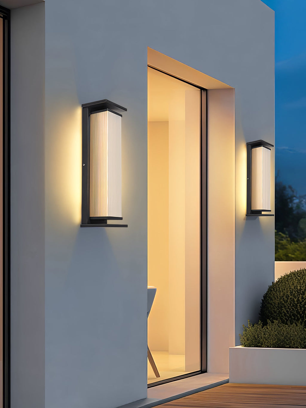Two Clérachic wall lights, one tall and one short, on a pale outside wall by a sliding glass door that opens onto a timber deck.