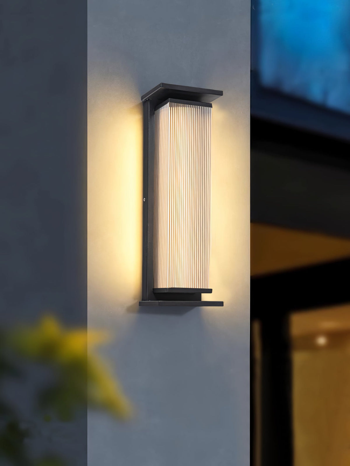 A Clérachic wall light lit warm on a grey wall beside a corner, with a blurred shrub in the foreground.