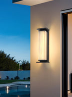 A Clérachic wall light beside a patio door at dusk, with a pool and trees behind it under a deep blue sky.