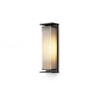 A mid-height Clérachic wall light with a black frame and a ribbed shade lit warm, on a plain white backdrop.