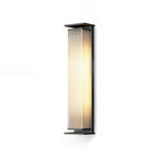 A tall Clérachic wall light with a ribbed shade lit warm, on a plain white backdrop and a soft shadow.