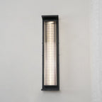 Front view of a tall Clérachic wall light in black, its slim ribbed shade lit with a bright white strip against a lace curtain.