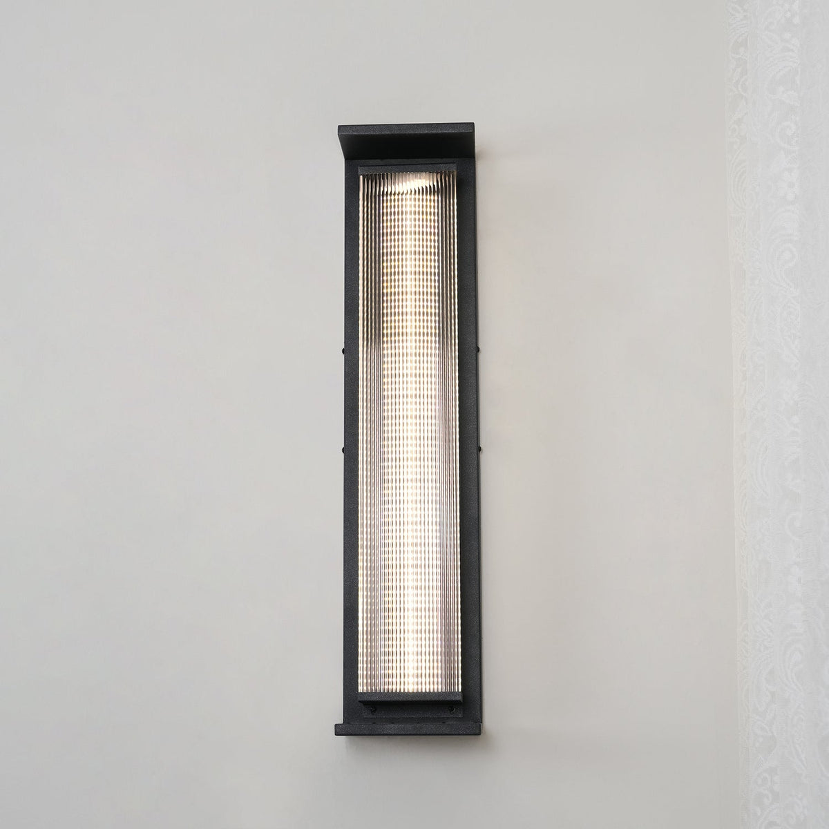 Front view of a tall Clérachic wall light in black, its slim ribbed shade lit with a bright white strip against a lace curtain.