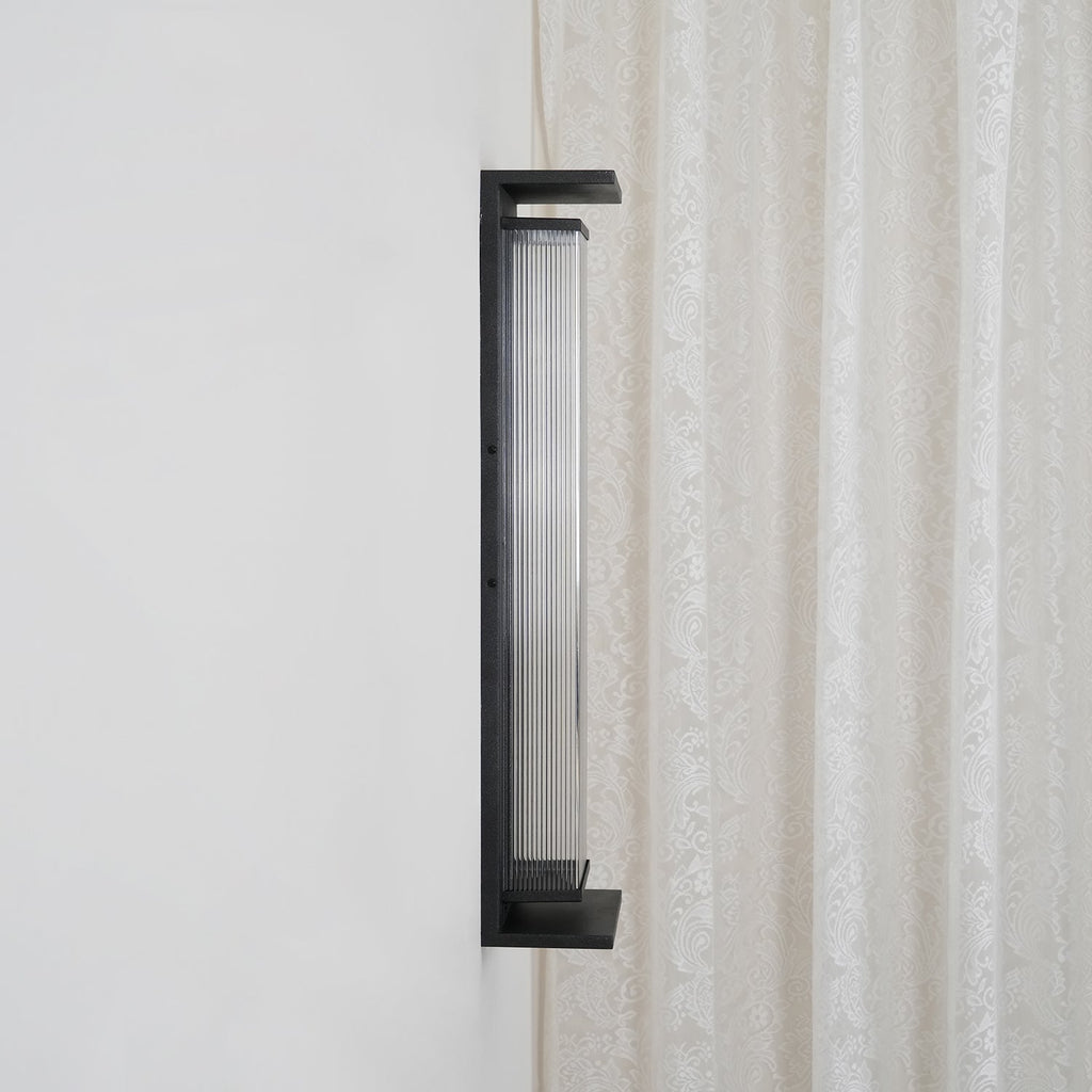 Side view of the same Clérachic wall light unlit, with a dark ribbed shade inside the black frame, beside a lace curtain.