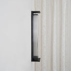 Side view of the same Clérachic wall light unlit, with a dark ribbed shade inside the black frame, beside a lace curtain.