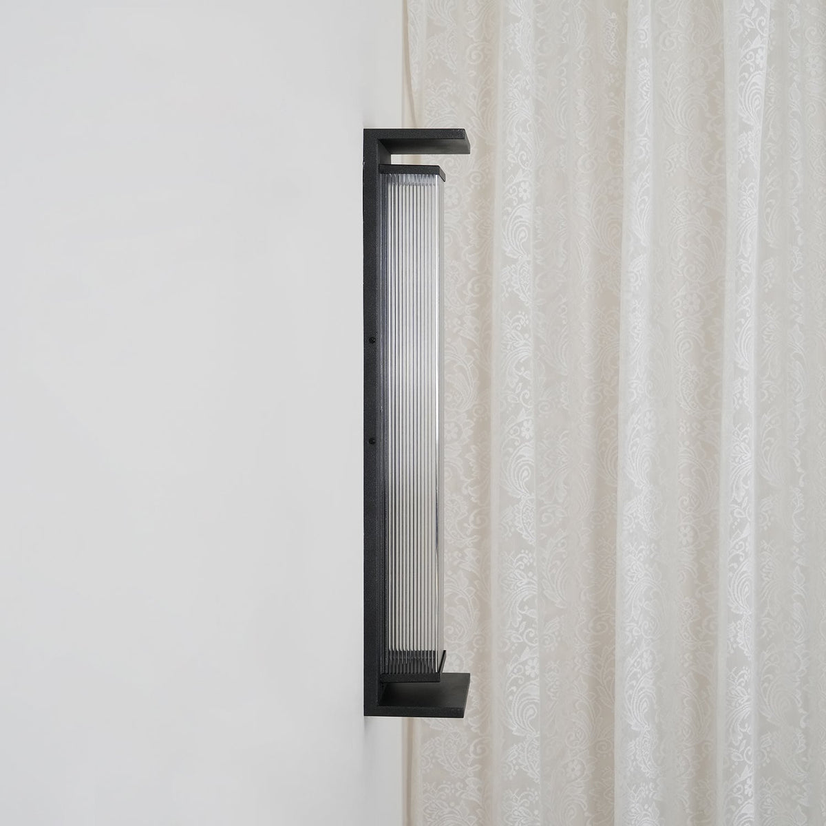 Side view of the same Clérachic wall light unlit, with a dark ribbed shade inside the black frame, beside a lace curtain.