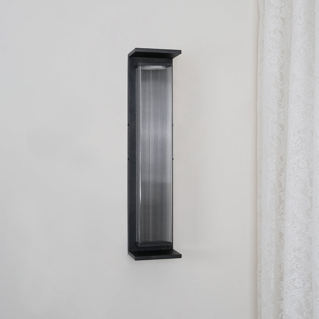 This Clérachic wall light unlit on a pale wall, its black frame and clear ribbed glass showing a grey tone.