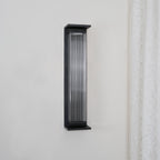 This Clérachic wall light unlit on a pale wall, its black frame and clear ribbed glass showing a grey tone.