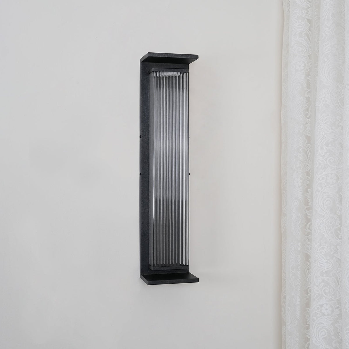 This Clérachic wall light unlit on a pale wall, its black frame and clear ribbed glass showing a grey tone.