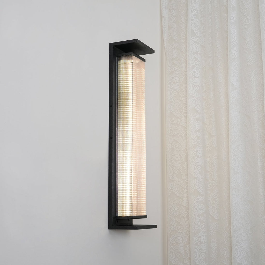 Front view of this Clérachic wall light lit warm white, with a pale strip of light behind the fluted glass in a black frame.