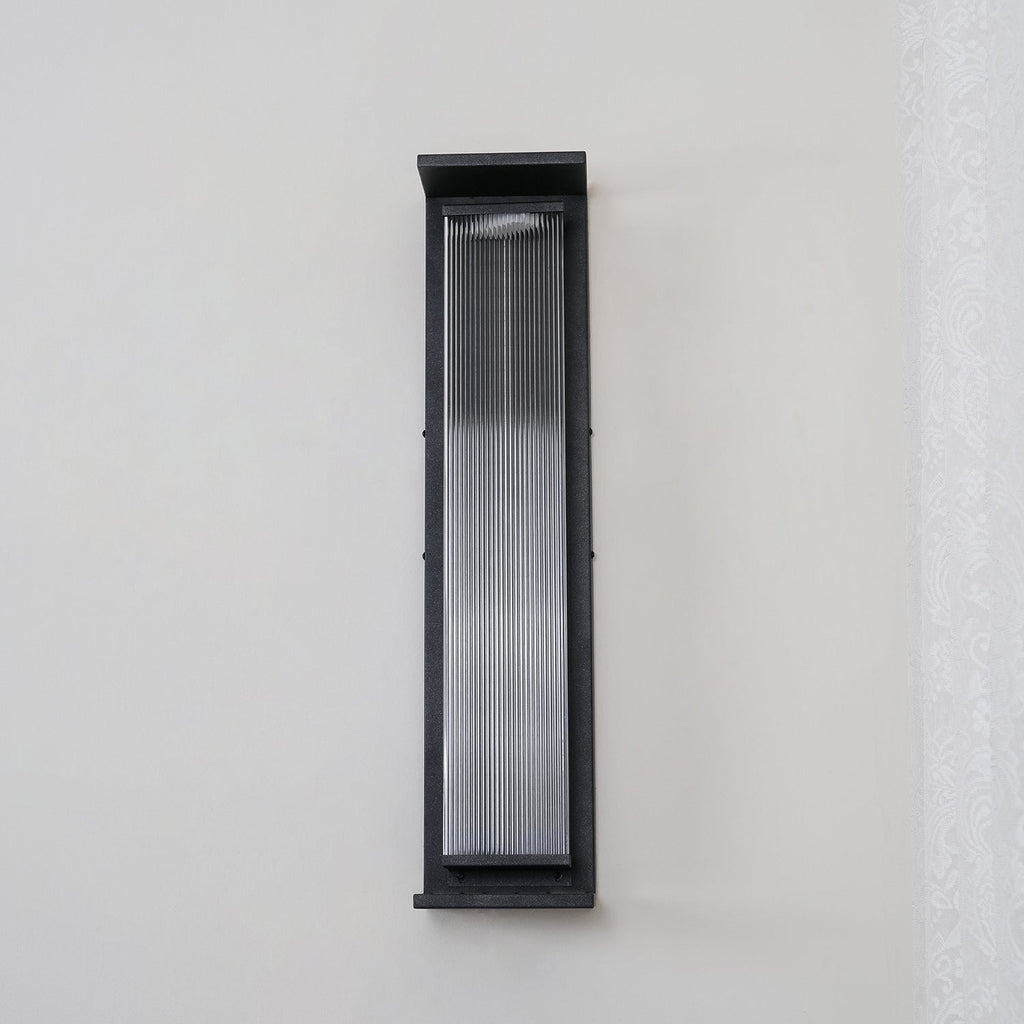 A Clérachic wall light unlit on a plain pale wall, with a black frame around clear ribbed glass.