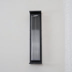 A Clérachic wall light unlit on a plain pale wall, with a black frame around clear ribbed glass.