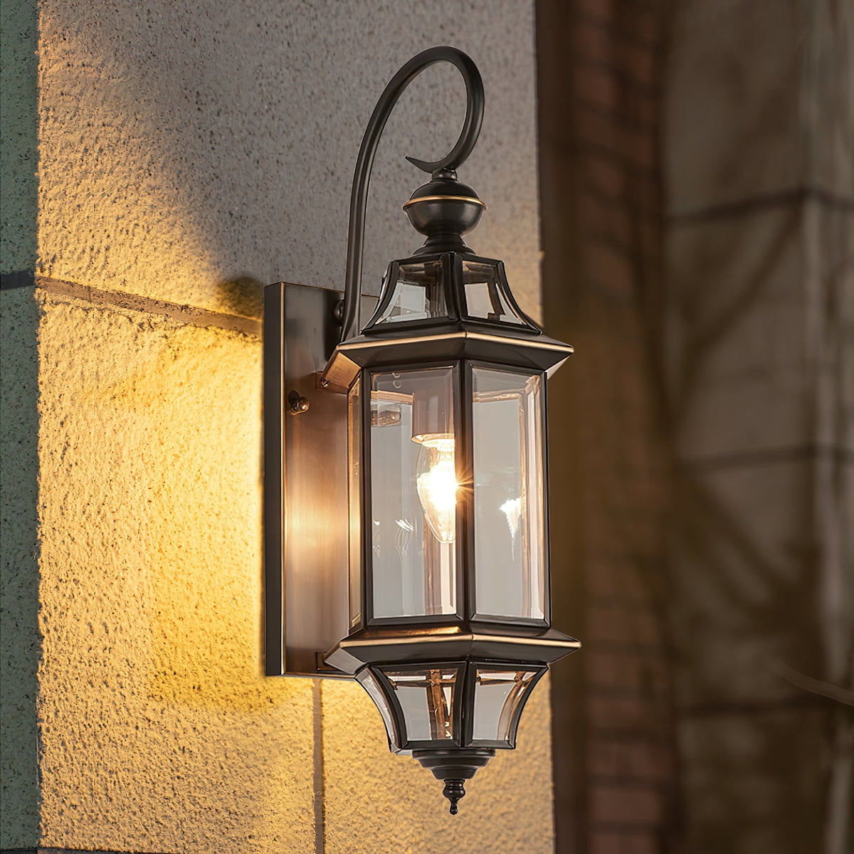Warm light glows through the clear glass of this Clérachic outdoor wall lantern, mounted on a rough yellow-lit stucco wall.