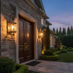 A pair of Clérachic wall lanterns lit at dusk beside a double wooden front door, on a stone-clad house front.